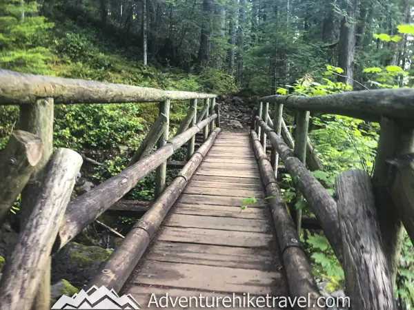 Trail to Upper Lena Lake. Soon after passing this moss-laden landscape you will cross some rustic wooden bridges through the woods. Next, you will come across a gigantic boulder that towers above the trail. There are several small boulders underneath this huge rock where people often sit to take a rest break and eat a snack. After the massive boulder, there is a larger wooden bridge going over a small stream.